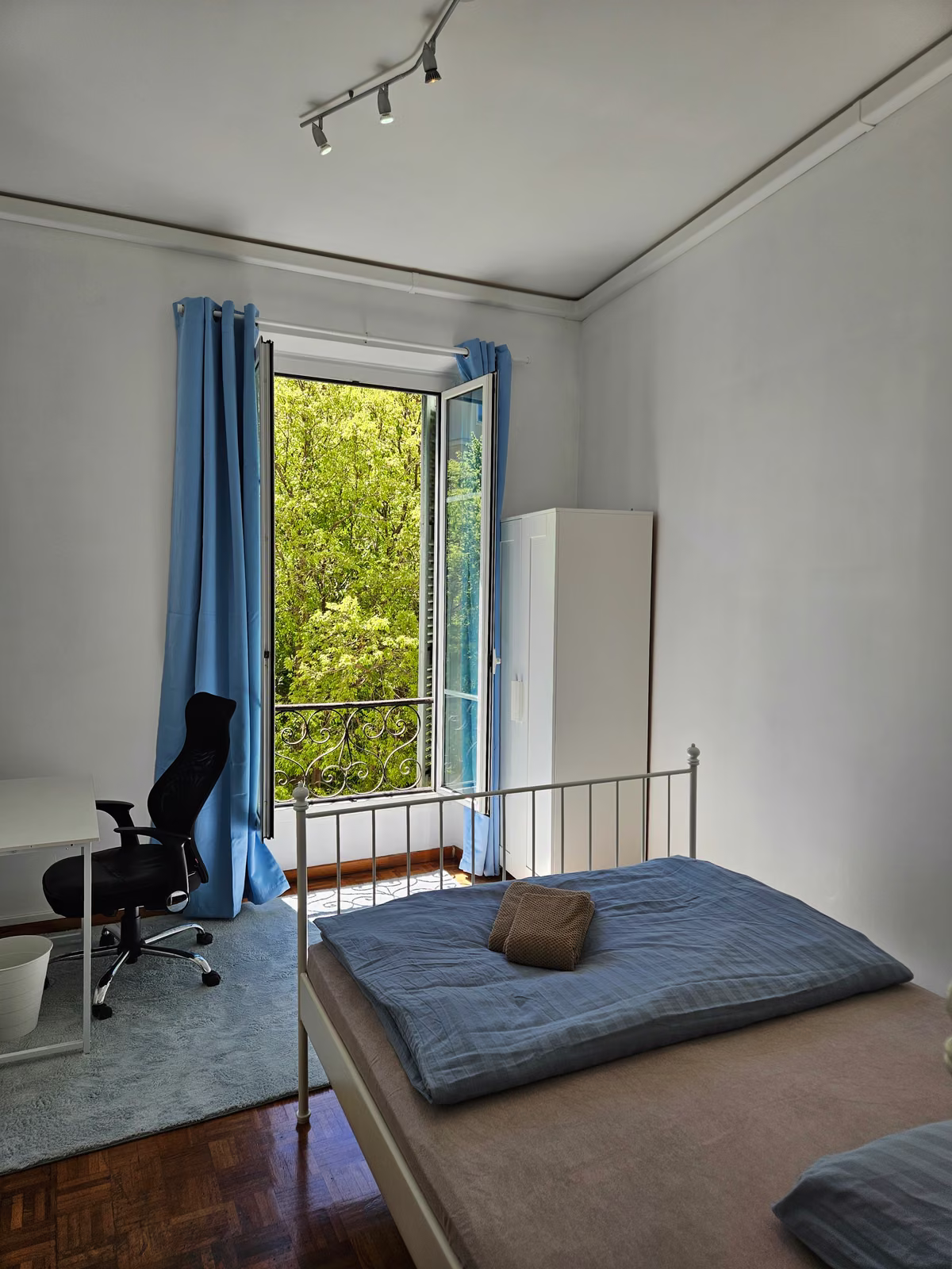 A double room with an office chair at the desk and the balcony doors open onto greenery
