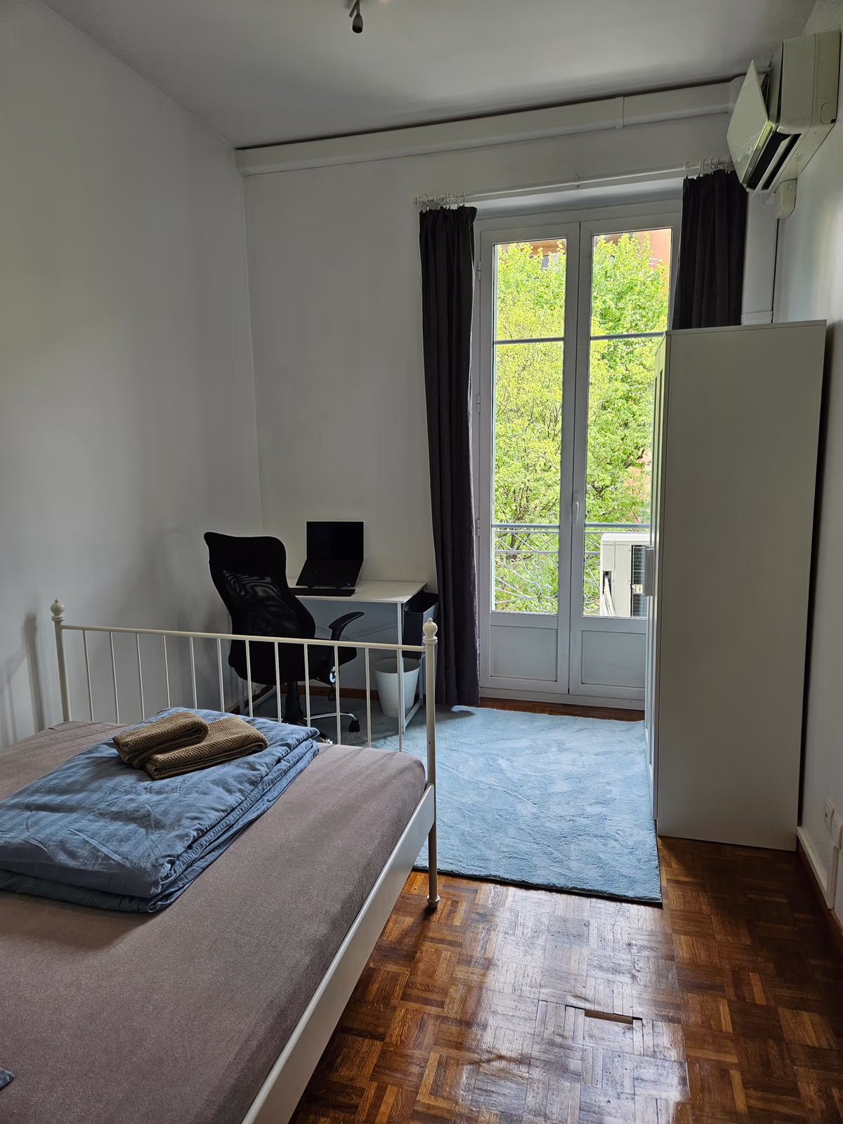 A double room with a white iron bed, a desk and a wardrobe by the balcony door
